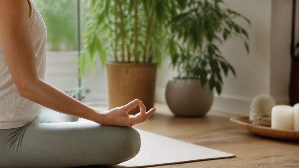 Personne en posture de méditation assise sur un tapis de yoga dans un intérieur lumineux et calme avec des plantes vertes